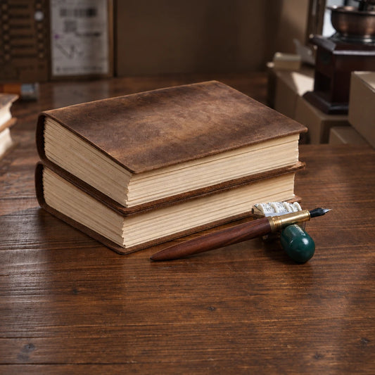 Two brown leather journals stacked on a wooden desk beside a wooden dip pen and green pen rest.