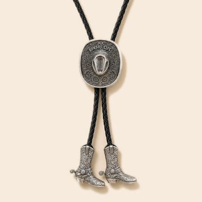 Silver Cowboy Life hat bolo tie with floral tooling and two cowboy boot pendants on a warm ivory background