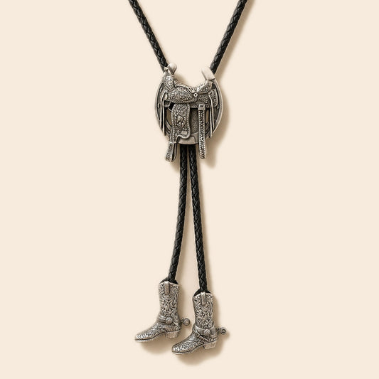 Silver Western saddle bolo tie with engraved tooling and two cowboy boot pendants on a warm ivory background