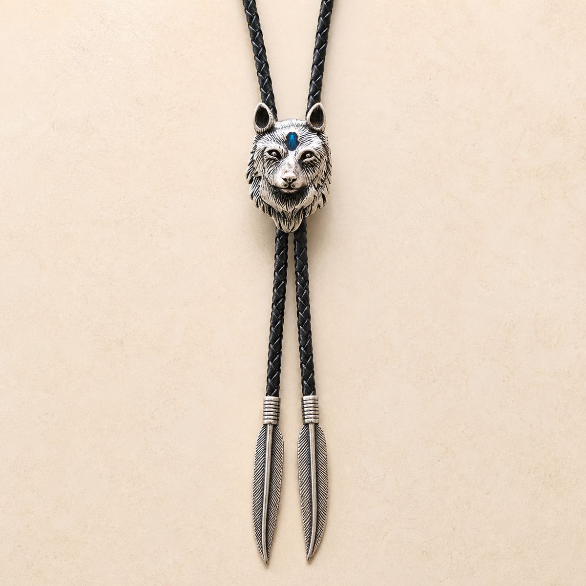 Stainless steel blue inlay wolf head bolo tie with black braided leather cord and feather-shaped tips on a warm ivory background