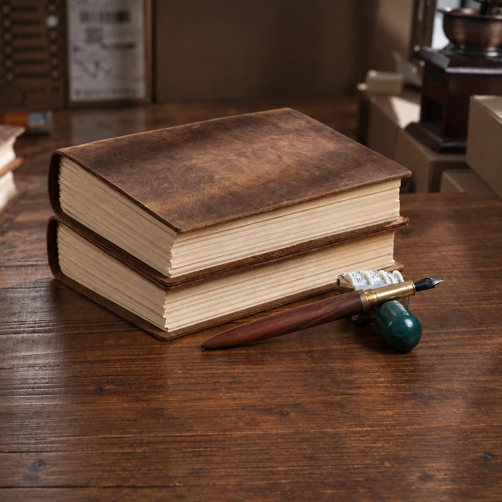 Two brown leather journals stacked on a wooden desk beside a wooden dip pen and green pen rest.