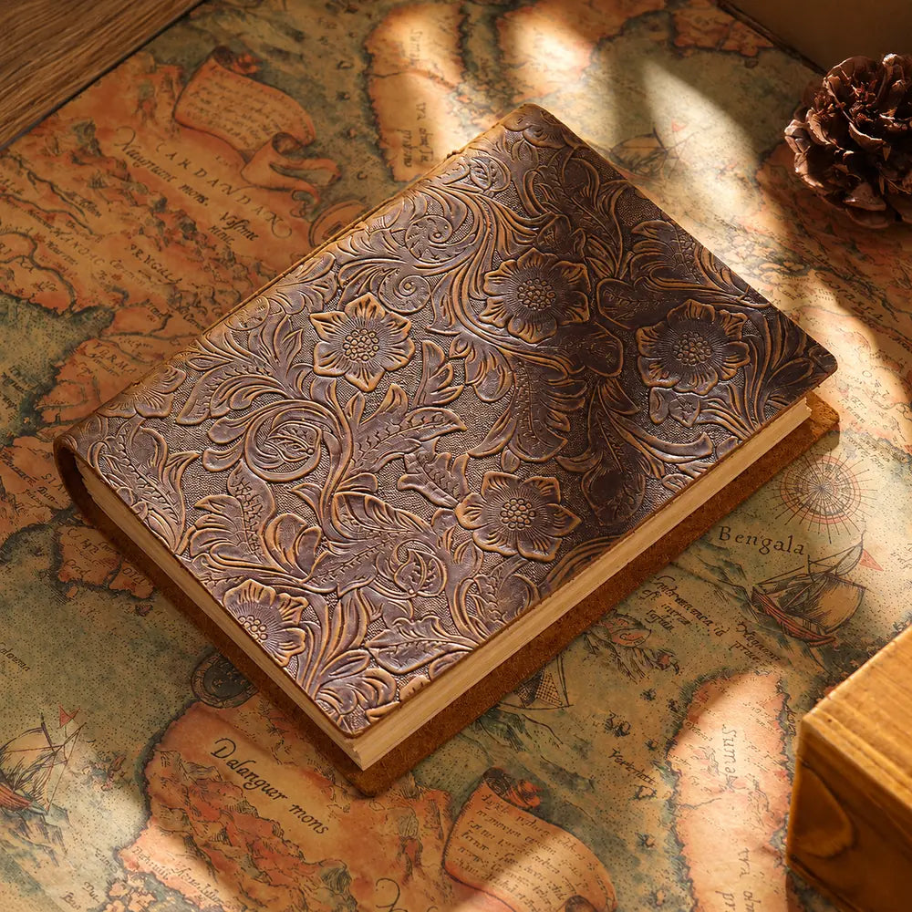 Floral-patterned brown leather journal on a vintage map in warm sunlight beside a pinecone.