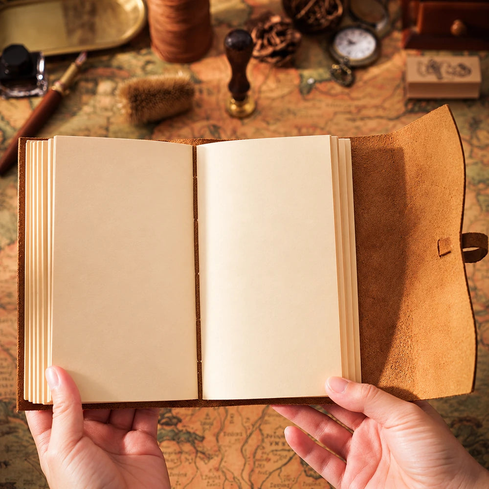Hands holding an open portrait leather journal with blank cream pages and a brown wraparound flap over a vintage map.